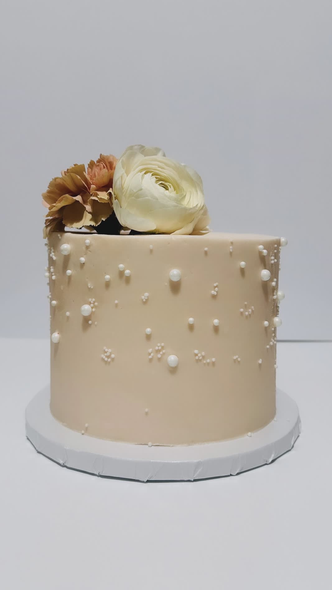 Elegant beige cake decorated with white pearls and fresh flowers, perfect for weddings or special events.