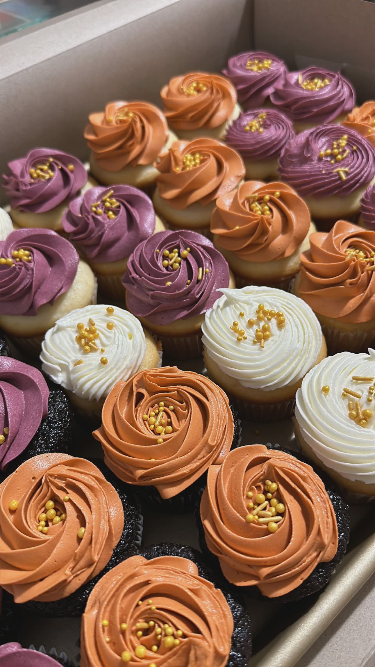 Box of assorted cupcakes with vibrant orange, purple, and white frosting, topped with golden sprinkles.